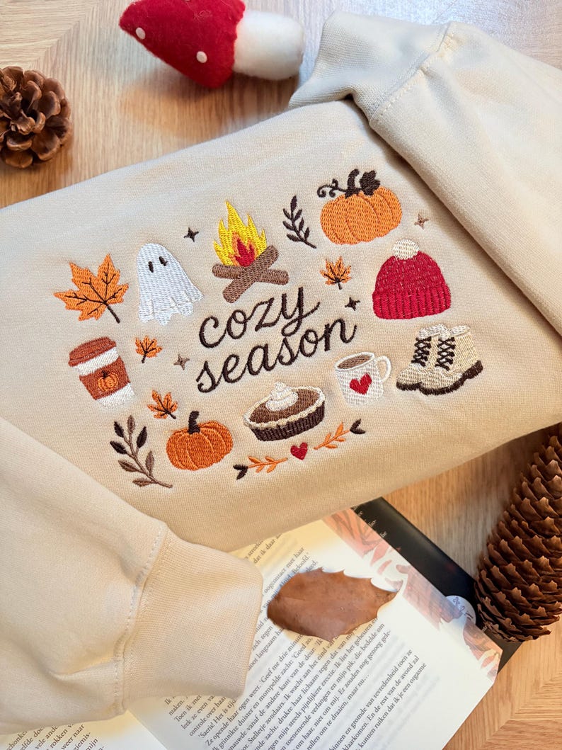 Geborduurde beige sweatshirt met het opschrift 'Cozy Season' en een herfstig borduurontwerp van een kampvuur, pompoenen, spookje, pumpkin spice koffie, warme chocolademelk, herfstbladeren, taart, gebreide muts en winterlaarsjes. De sweater ligt op een open boek met dennenappels, kaarslicht en herfstdecoratie. Perfecte herfsttrui voor Halloween, cozy season en herfstliefhebbers.