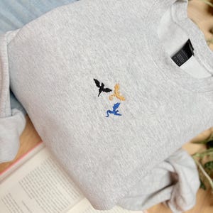 Embroidered dragon book sweatshirt for fantasy book lovers, cozy bookish dragon crewneck with detailed dragon embroidery. Perfect gift for readers, BookTok fans, librarians and fantasy readers. Aesthetic book lover sweatshirt inspired by dragons and magical stories, ideal bookish merch for reading lovers, dragon readers and fantasy literature fans.