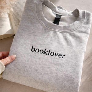 Puede incluir: Una sudadera gris claro con la palabra "booklover" bordada en negro. La sudadera doblada está sobre una superficie con un libro y una planta decorativa visibles. La sudadera de cuello redondo es de estilo casual.