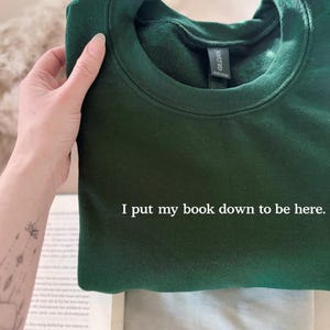 May include: A dark green sweatshirt with the text "I put my book down to be here." The sweatshirt is folded and held by a hand. The Gildan brand label is visible. A book is partially visible in the background.