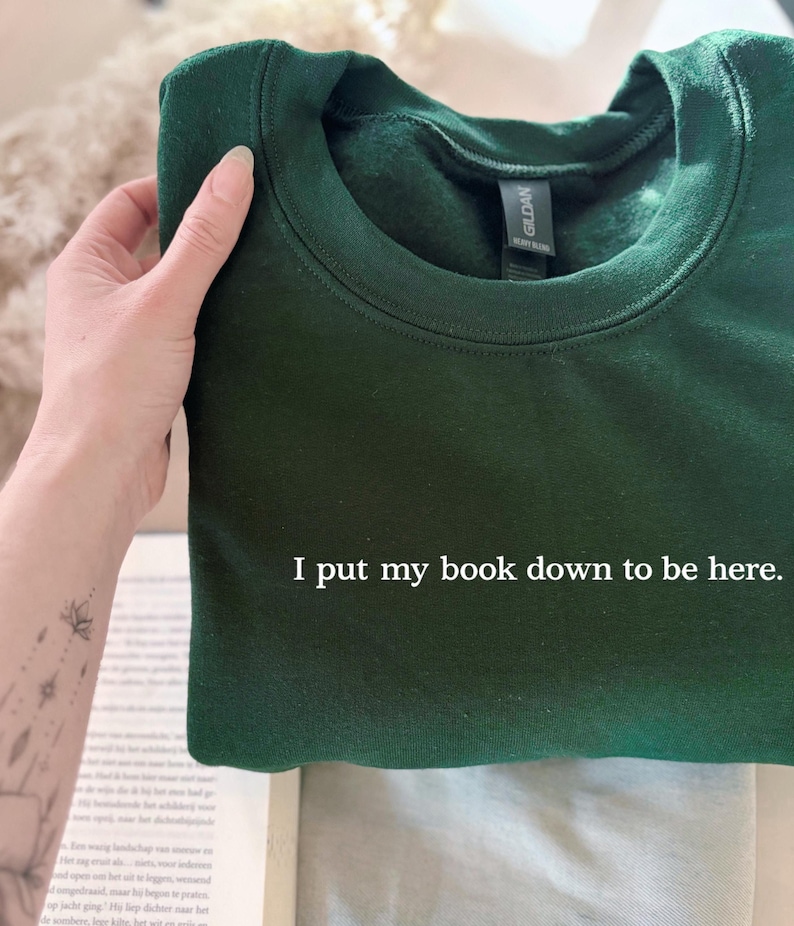 May include: A dark green sweatshirt with the text "I put my book down to be here." The sweatshirt is folded and held by a hand. The Gildan brand label is visible. A book is partially visible in the background.