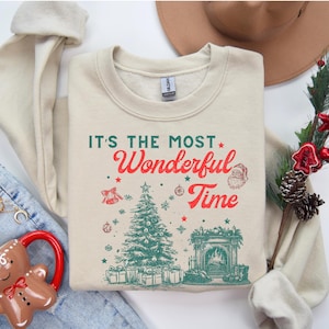 Può includere: Felpa color crema con la scritta "IT'S THE MOST Wonderful Time" in rosso e verde. Il design include un albero di Natale, un camino e Babbo Natale. Un capo festivo a tema natalizio.