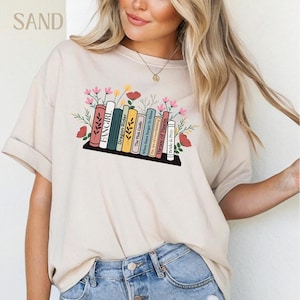 Op de afbeelding: Zandkleurig T-shirt met een boekenplankontwerp met kleurrijke boekruggen en bloemaccenten. De boeken bevatten titels als "FANGIRL" en "Throne of Thorns". Het woord "SAND" staat linksboven gedrukt. Casual stijl.