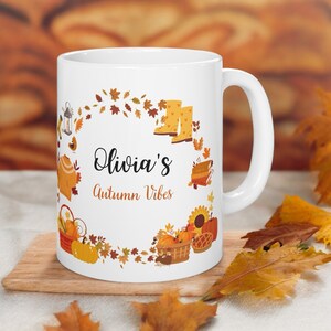 Puede incluir: Taza de cerámica blanca con un diseño de otoño. La taza presenta una corona de hojas de otoño, calabazas, un pastel, un girasol, una linterna, un suéter y botas de lluvia. El texto "Olivia's Autumn Vibes" está impreso en la taza.