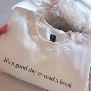 Può includere: Felpa color crema con la scritta "It's a good day to read a book" in lettere marroni. La felpa ha un girocollo e maniche lunghe. Nel'immagine sono visibili anche un libro e piante secche.