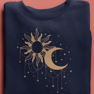 Puede incluir: Sudadera azul marino con un diseño dorado de sol y luna creciente con estrellas colgantes. El gráfico celestial está centrado en la parte delantera de la prenda doblada. La sudadera está hecha de un material suave.