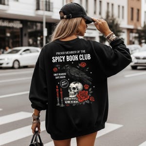 May include: Black sweatshirt with the text "PROUD MEMBER OF THE SPICY BOOK CLUB." The graphic includes a raven, skull, roses, and candles. "SMUT READERS' SAFE HAVEN" and "MEET WEEKLY" are also printed on the sweatshirt.
