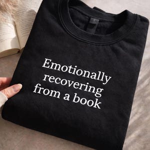 Pode incluir: Camisola preta de gola redonda com o texto branco "Emotionally recovering from a book". A camisola está dobrada, com um livro e uma planta decorativa no fundo.
