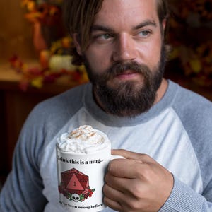 May include: White ceramic mug with the text "I think this is a mug.. we've been wrong before" and a red D20 die graphic. The mug is filled with a whipped topping and cinnamon. The mug is held by a person wearing a gray and white raglan shirt.