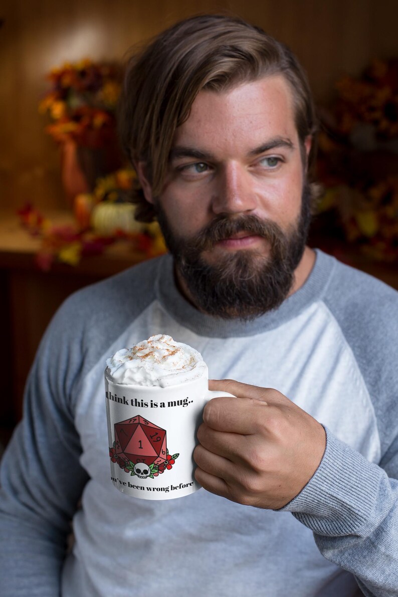 May include: White ceramic mug with the text "I think this is a mug.. we've been wrong before" and a red D20 die graphic. The mug is filled with a whipped topping and cinnamon. The mug is held by a person wearing a gray and white raglan shirt.