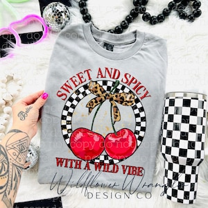 Op de afbeelding: Een grijs t-shirt met een afbeelding van twee rode kersen, een luipaardprint strik en de tekst "Sweet and Spicy With a Wild Vibe". Een zwart-wit geruite beker staat naast het shirt.