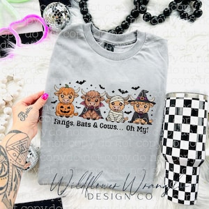 May include: A light gray t-shirt with a Halloween-themed graphic featuring four cartoon Highland cows dressed as a pumpkin, vampire, mummy, and witch. The text "Fangs, Bats & Cows... Oh My!" is below the cows. A black and white checkered tumbler is on the right.