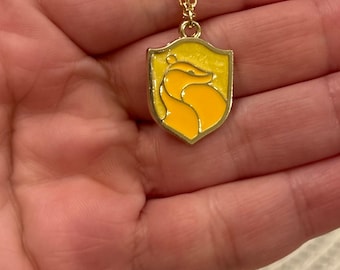May include: A gold-colored necklace with a pendant featuring a yellow shield with a badger design. The badger is in a lighter shade of yellow. The necklace is held in a hand.