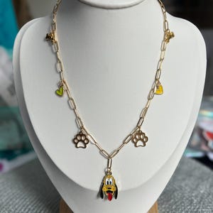 Puede incluir: Un collar de cadena dorada con un dije de un perro de dibujos animados con la lengua roja, dos dijes de huella de pata y dos dijes de corazón. Los dijes de corazón son verdes y amarillos.