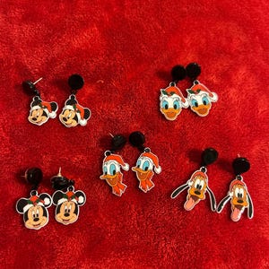May include: A collection of six pairs of Christmas-themed earrings featuring cartoon characters. The earrings showcase Mickey Mouse, Minnie Mouse, Donald Duck, and Pluto, all wearing Santa hats. The earrings have black posts and are set against a red background.