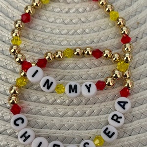 Könnte beinhalten: Drei Armbänder mit Perlen. Die Armbänder bestehen aus goldfarbenen Perlen, roten und gelben facettierten Perlen und weißen Buchstabenperlen. Die Buchstabenperlen schreiben "CHIEFS" und "IN MY ERA".