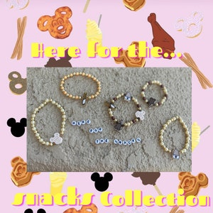May include: Four gold beaded bracelets with different Disney-themed charms. The charms include a Mickey Mouse head, a Mickey Mouse pretzel, a Mickey Mouse ice cream cone, and a Mickey Mouse waffle. The bracelets are arranged on a light brown surface. The text "Here for the... Snacks Collection" is written in yellow letters on a pink background.