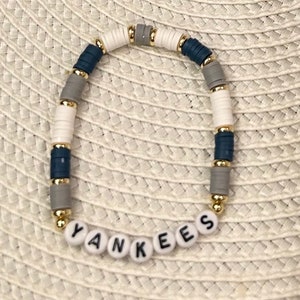 Op de afbeelding: Een witte, grijze en blauwe kralen armband met gouden accenten en het woord "YANKEES" gespeld in witte letters.