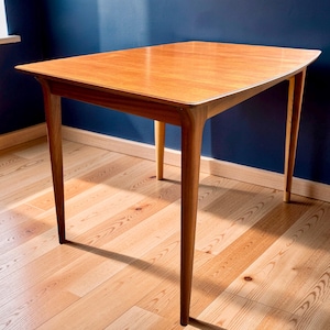 May include: A rectangular wooden table with a smooth, light brown finish and a visible wood grain pattern. The table features four tapered legs that extend outwards from the corners, supporting a flat top with slightly rounded edges.