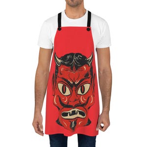May include: Red apron with a black and red devil face illustration. The apron has adjustable black straps and two black buttons.