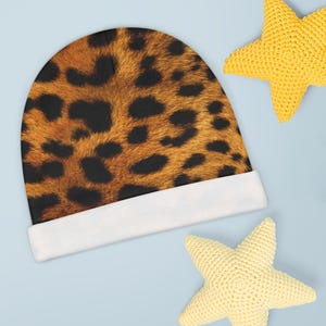 May include: A baby beanie hat with a leopard print design, featuring brown and black spots on a tan background. The hat has a white cuff. Two yellow star-shaped toys are in the background.