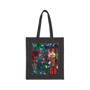 May include: Black tote bag with a Christmas-themed design. The image shows a detailed nutcracker soldier figurine with a gold staff, red trousers, and a blue jacket, set against a backdrop of a decorated Christmas tree with lights and ornaments.