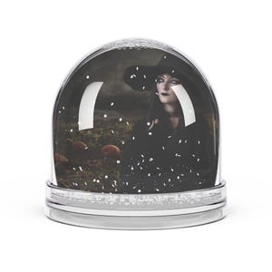 Peut inclure: Une boule à neige en verre transparent contenant une scène sombre et atmosphérique d'une personne portant un chapeau noir à larges bords et des vêtements sombres dans un champ de citrouilles, avec des paillettes blanches imitant la neige.