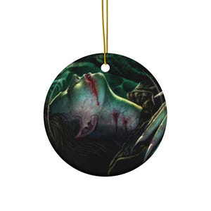 Puede incluir: Un adorno de cerámica con una ilustración en verde oscuro y negro de una persona con sangre goteando de su boca y heridas en el cuello. La persona está acostada de espaldas con los ojos cerrados.