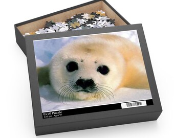 Puede incluir: Caja de rompecabezas con una imagen de una cría de foca. La foca tiene pelaje blanco cremoso y grandes ojos negros. La caja está abierta, mostrando las piezas del rompecabezas. La caja es negra y tiene el texto "8x10 Puzzle 120 pcs Ages 9+".