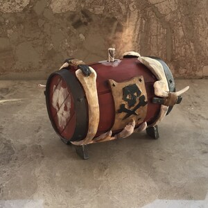 May include: A red wooden barrel with a skull and crossbones design on a metal shield. The barrel is decorated with bone-like accents and has a metal band around the middle. The barrel is sitting on a concrete surface.