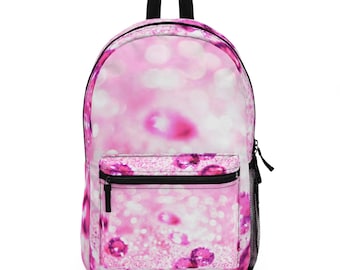 May include: Pink backpack with a glittery, bokeh-style design. It has a front zipped pocket, a black zip, a black top handle, and a mesh side pocket. The design is a mix of pink and white, suitable for school or travel.