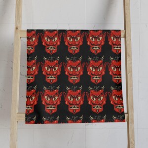 May include: A black and red patterned towel featuring a repeating design of a devil's face. The devil's face is red with yellow eyes, black hair, and horns. The towel is hanging on a wooden rack.