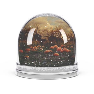 May include: A clear glass snow globe with a silver base. Inside, a fall scene with pumpkins and bare trees under a cloudy sky. White glitter simulates falling snow, creating a seasonal decorative item.