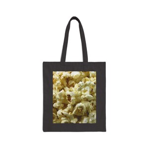 May include: Black tote bag featuring a close-up image of freshly popped popcorn. The popcorn is a mix of white and yellow kernels, with a textured appearance. The bag has a rectangular design with a black handle.