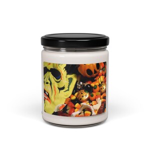 Op de afbeelding: Een witte kaarsenpot met een zwart deksel. Het etiket heeft een Halloween-thema met een zombiegelaat, pompoenen, snoepmaïs en andere feestelijke elementen. De pot is gevuld met was.