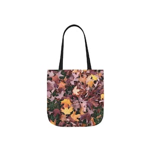 May include: Tote bag featuring a vibrant autumn leaf design. The bag's pattern showcases a mix of brown, orange, and yellow leaves scattered across a green background. It has black handles and is perfect for carrying groceries or personal items.