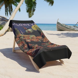 Peut inclure: Une serviette de plage sur le thème d'Halloween, posée sur une chaise longue en bois. La serviette présente une illustration vintage avec des citrouilles grimaçantes, une sorcière et le mot "HALLOWEEN" en blanc sur fond sombre.