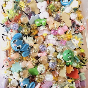 May include: A vibrant collection of small charms and pendants in diverse shapes and designs. The assortment features cartoon characters, glitter-filled globes, and food-themed items in blue, pink, yellow, and green. Many have small metal loops for attaching to jewellery or other crafts.