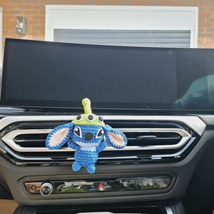 Könnte beinhalten: Ein blaues, weißes und grünes gehäkeltes Charakter mit großen Ohren und einem Hut, der an der Lüftungsöffnung eines Autos befestigt ist. Die Figur ist eine beliebte Zeichentrickfigur. Der Innenraum des Autos verfügt über einen großen Bildschirm und Lüftungsschlitze.