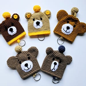 May include: Five handmade crochet bear keyrings in shades of brown, tan, and yellow. Each keyring features a bear face with a white muzzle, black eyes, and a smiling mouth. The keyrings have a small metal ring attached.