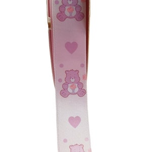 K&ouml;nnte beinhalten: Hellrosa Band mit sich wiederholenden Mustern von lila Care Bears, die Blumen halten, unterbrochen von rosa Herzen und Punkten. Dieses Band wird wahrscheinlich f&uuml;r Bastelarbeiten oder Geschenkverpackungen verwendet.