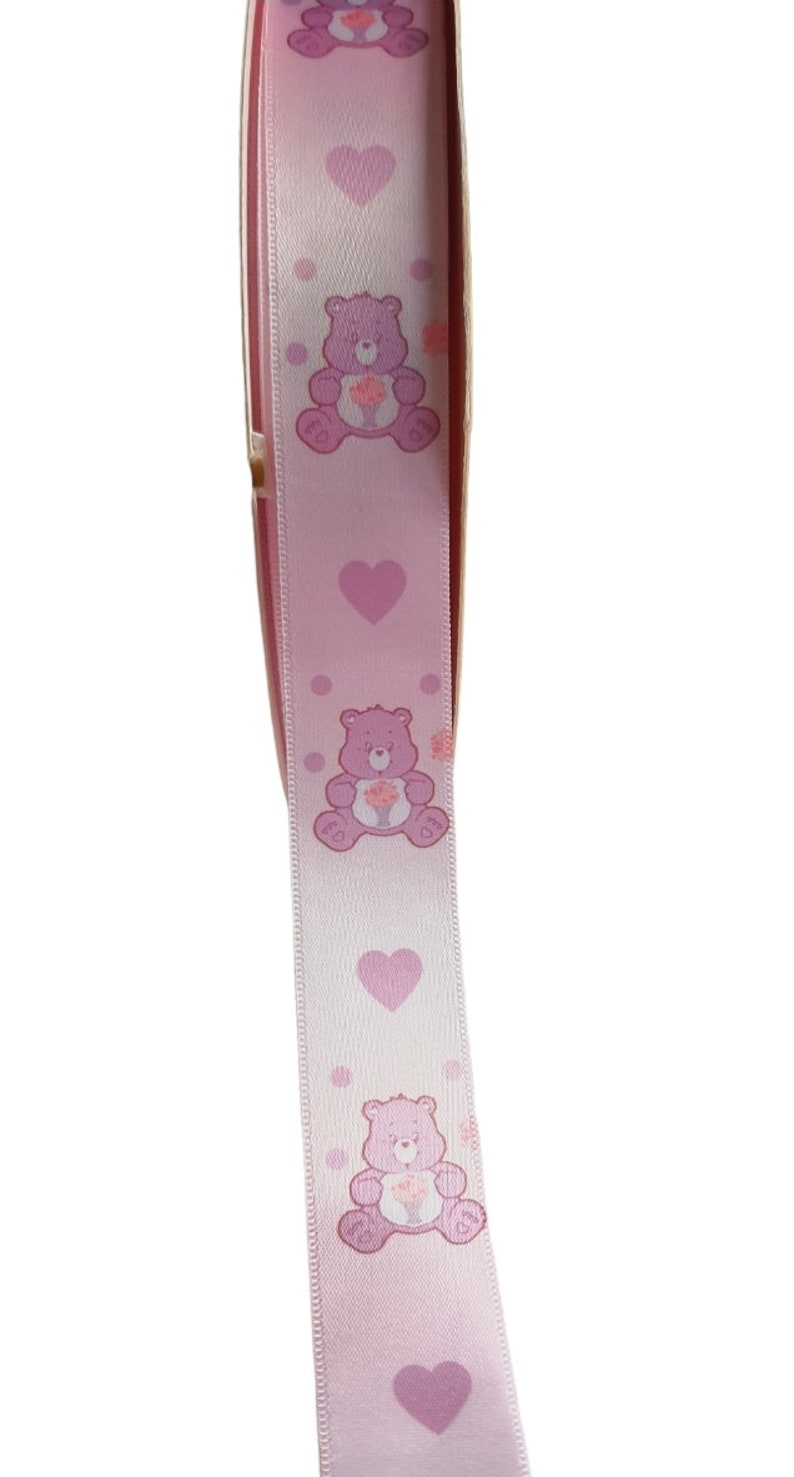 K&ouml;nnte beinhalten: Hellrosa Band mit sich wiederholenden Mustern von lila Care Bears, die Blumen halten, unterbrochen von rosa Herzen und Punkten. Dieses Band wird wahrscheinlich f&uuml;r Bastelarbeiten oder Geschenkverpackungen verwendet.