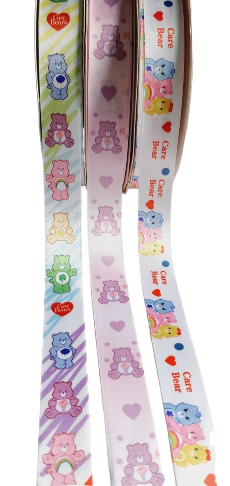 K&ouml;nnte beinhalten: Drei Rollen Band mit Care Bears-Designs. Die B&auml;nder zeigen verschiedene Care Bears-Charaktere, Herzen und Regenbogen. Die B&auml;nder sind in Pastellfarben gehalten, darunter Rosa, Blau, Gelb und Gr&uuml;n. Der Text "Care Bears" ist auf den B&auml;ndern aufgedruckt.