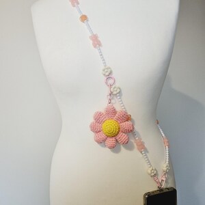 Könnte beinhalten: Ein Handyband mit einer rosa und gelben Häkelblume und einem Perlenband. Das Band hat weiße Perlen, rosa bärenförmige Perlen und einen rosa Verschluss. Das Telefon befindet sich in einer schwarzen Hülle.