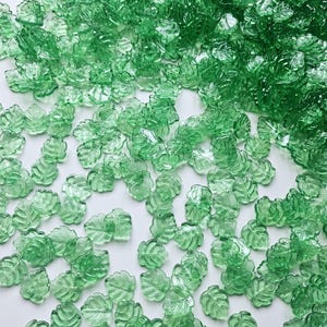 May include: A scattering of translucent green leaf-shaped beads on a white surface. The beads feature a detailed leaf vein design and vary in size. Suitable for jewellery making or craft projects.