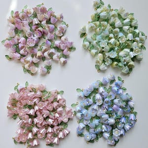 Könnte beinhalten: Vier Gruppen schillernder Blumenperlen in Rosa, Lila, Blau und Grün. Jede Gruppe zeigt zarte glockenförmige Blüten mit grünen Blättern und goldenen Akzenten, ideal für die Schmuckherstellung oder Bastelprojekte.