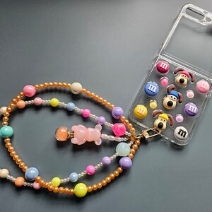 Könnte beinhalten: Eine transparente Handyhülle mit Cartoon-Hund-Charakteren und M&M's-Bonbons. Ein Perlen-Handyband in Pastellfarben, darunter Rosa, Gelb und Blau, ist mit einem goldenen Verschluss an der Hülle befestigt. Das Band hat einen kleinen rosa Bärenanhänger.