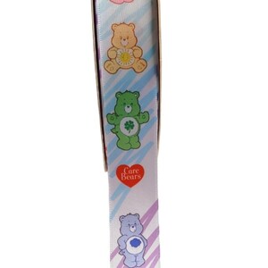 K&ouml;nnte beinhalten: Wei&szlig;es Band mit bunten Care Bears-Charakteren in verschiedenen Posen. Das Band hat ein diagonales Streifenmuster in Blau-, Lila- und Gr&uuml;nt&ouml;nen. Ein rotes Herz mit der Aufschrift "Care Bears" ist ebenfalls sichtbar.