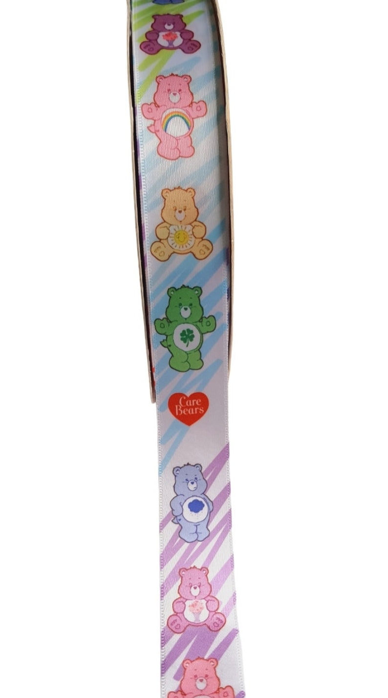 K&ouml;nnte beinhalten: Wei&szlig;es Band mit bunten Care Bears-Charakteren in verschiedenen Posen. Das Band hat ein diagonales Streifenmuster in Blau-, Lila- und Gr&uuml;nt&ouml;nen. Ein rotes Herz mit der Aufschrift "Care Bears" ist ebenfalls sichtbar.