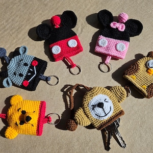 May include: A selection of handmade crochet keyrings in various character designs. The keyrings feature Mickey Mouse, Minnie Mouse, a bear, an owl, and other cartoon characters. Each keyring has a metal ring for keys and a pom-pom.